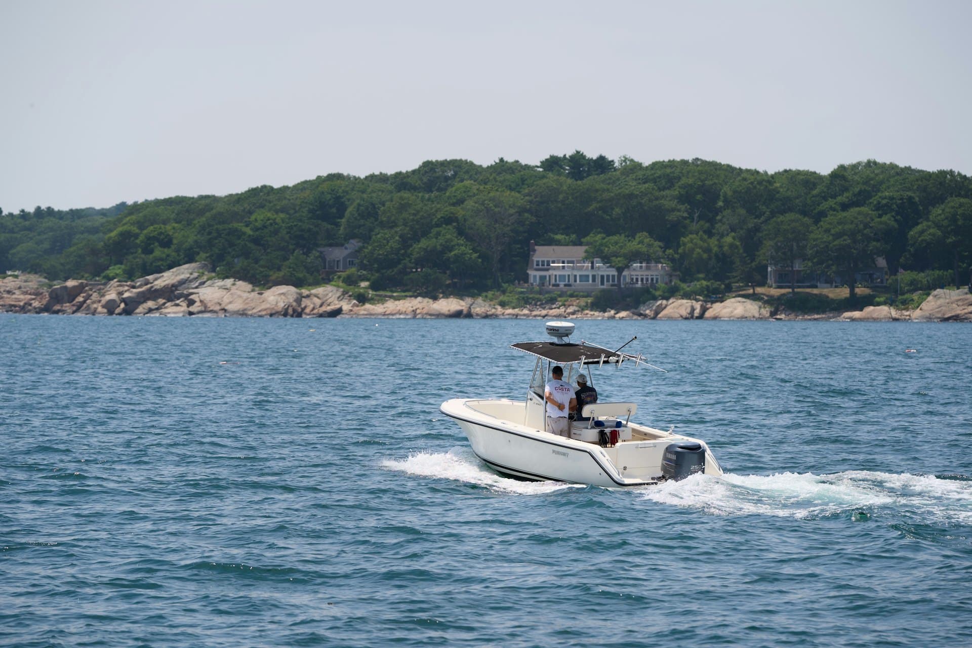 A centre-console boat with a T-top running past a wooded, rocky shoreline