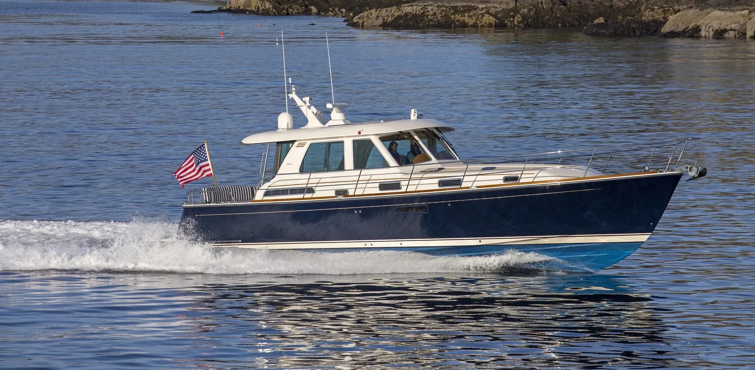 A navy-hulled Downeast cruiser with a teak-trimmed hardtop running past a rocky shore, its wake behind it