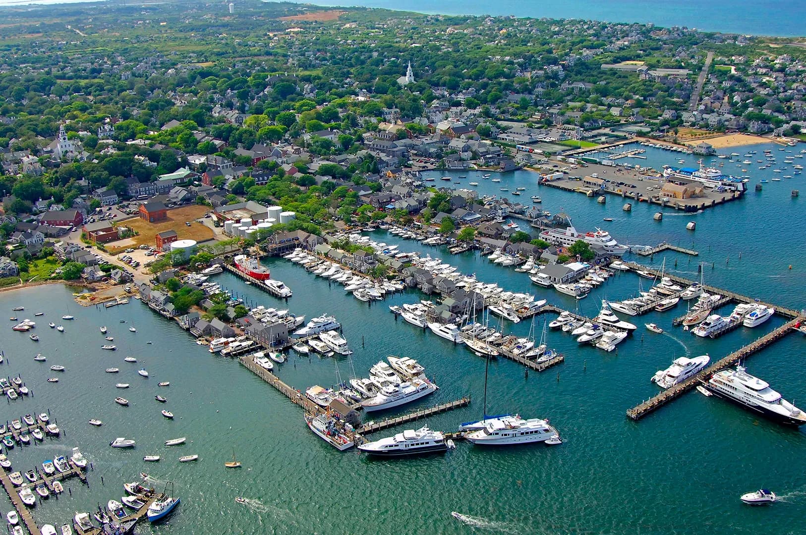 An island harbour town from the air, yachts berthed along its piers and the open sea beyond